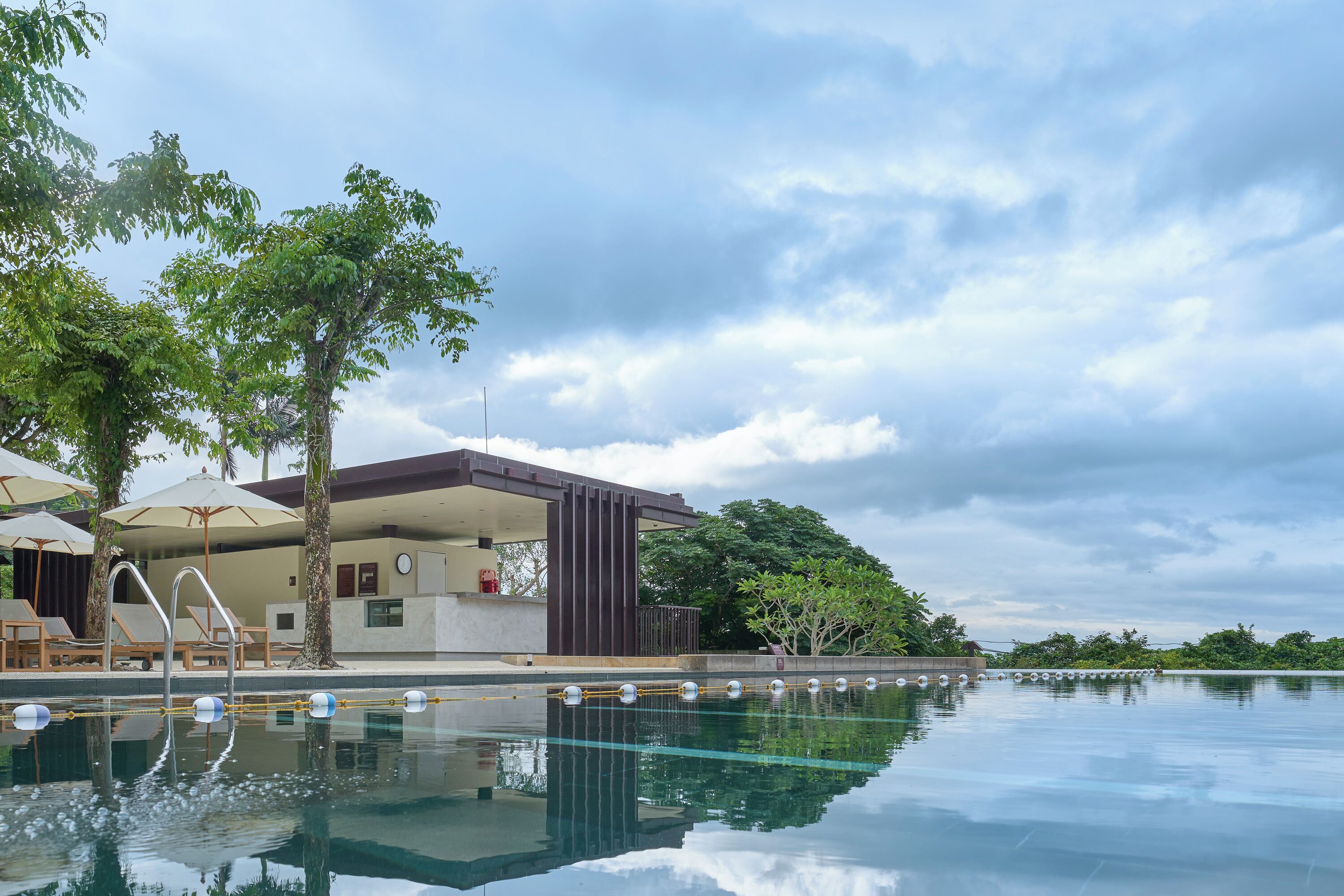 A swimming pool with lounge chairs and umbrellas, a modern building with a poolside area, and a cloudy sky.