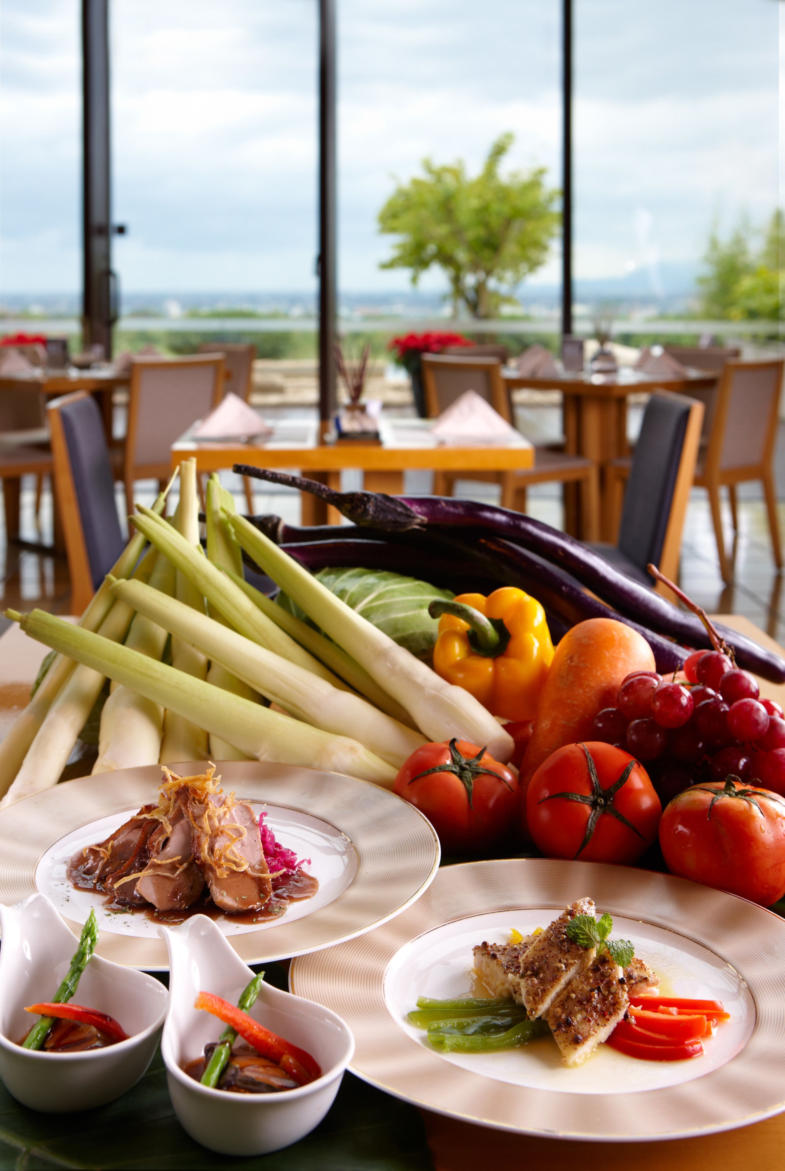 A dining table with various dishes and fresh vegetables, set in a room with large windows and a view of the outdoors.