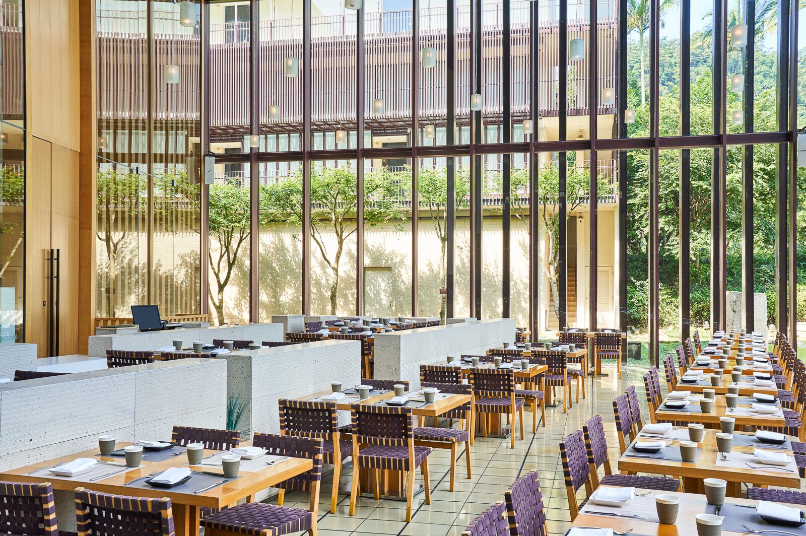 A spacious indoor dining area with long tables and chairs, large windows, and a view of greenery outside.