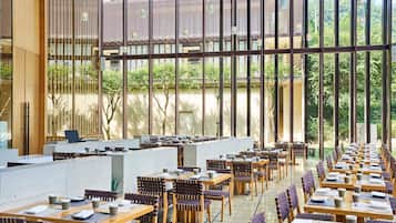 A spacious indoor dining area with long tables and chairs, large windows, and a view of greenery outside.