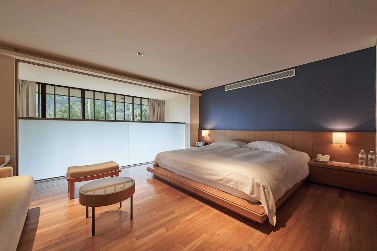 Un dormitorio moderno con una cama grande, mesitas de noche, un pequeño rincón de descanso y un amplio ventanal con vistas a zonas verdes.