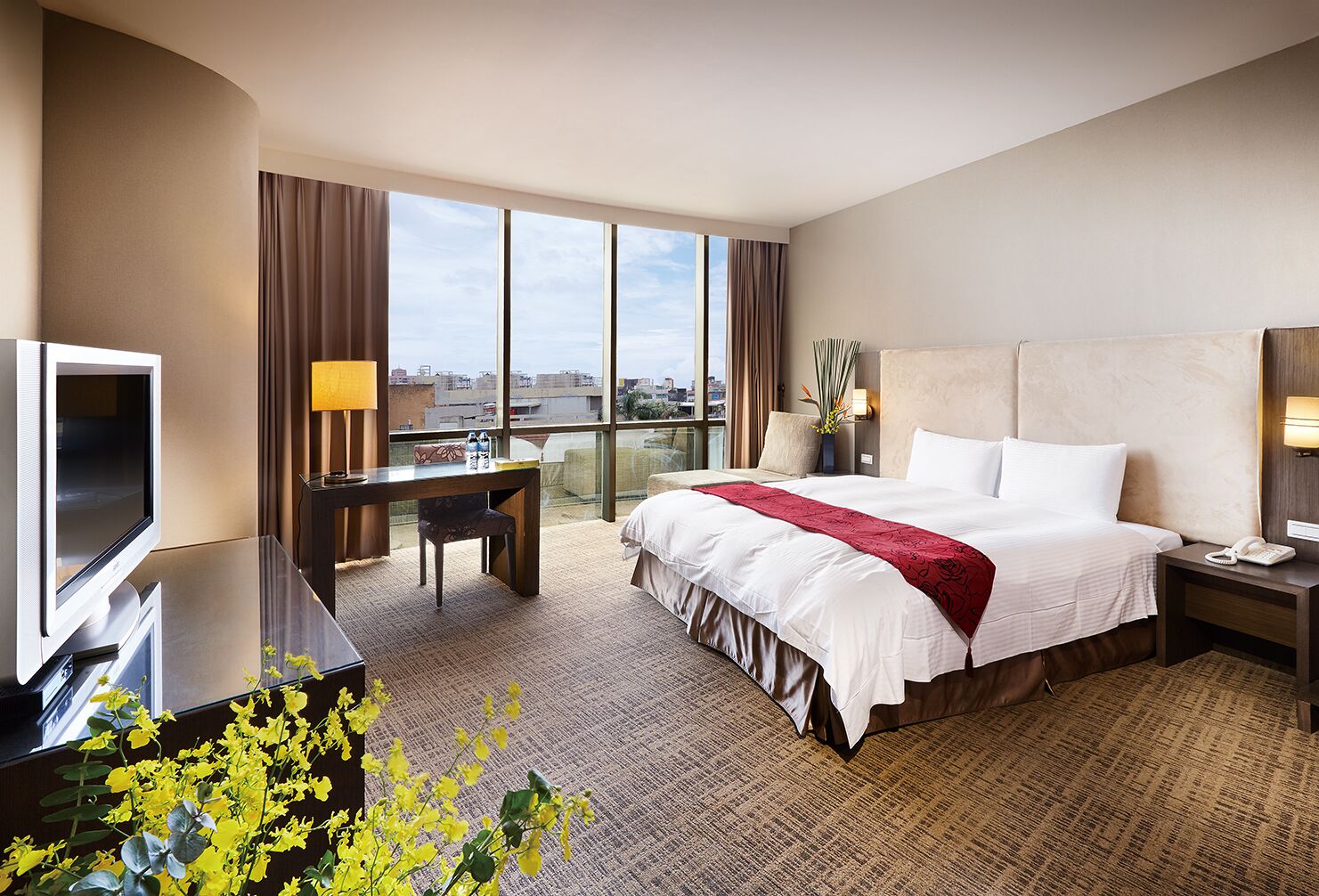 A hotel room with a large bed, a desk with a chair, a TV, and a view of the city through large windows.