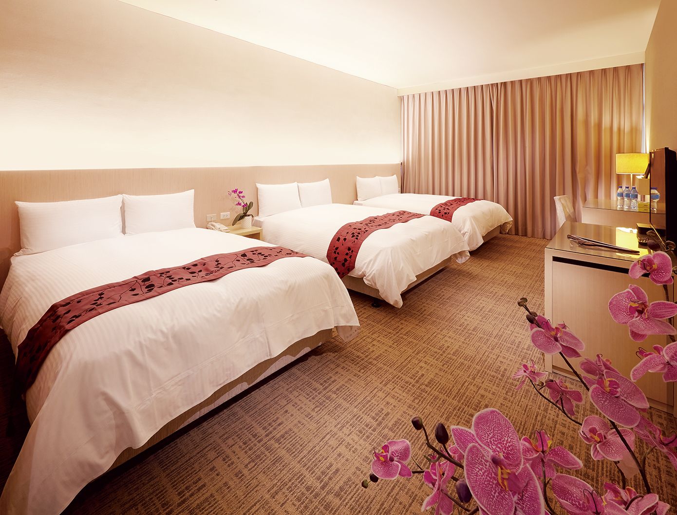 A hotel room with two beds, a desk, a chair, a lamp, and a decorative orchid plant.