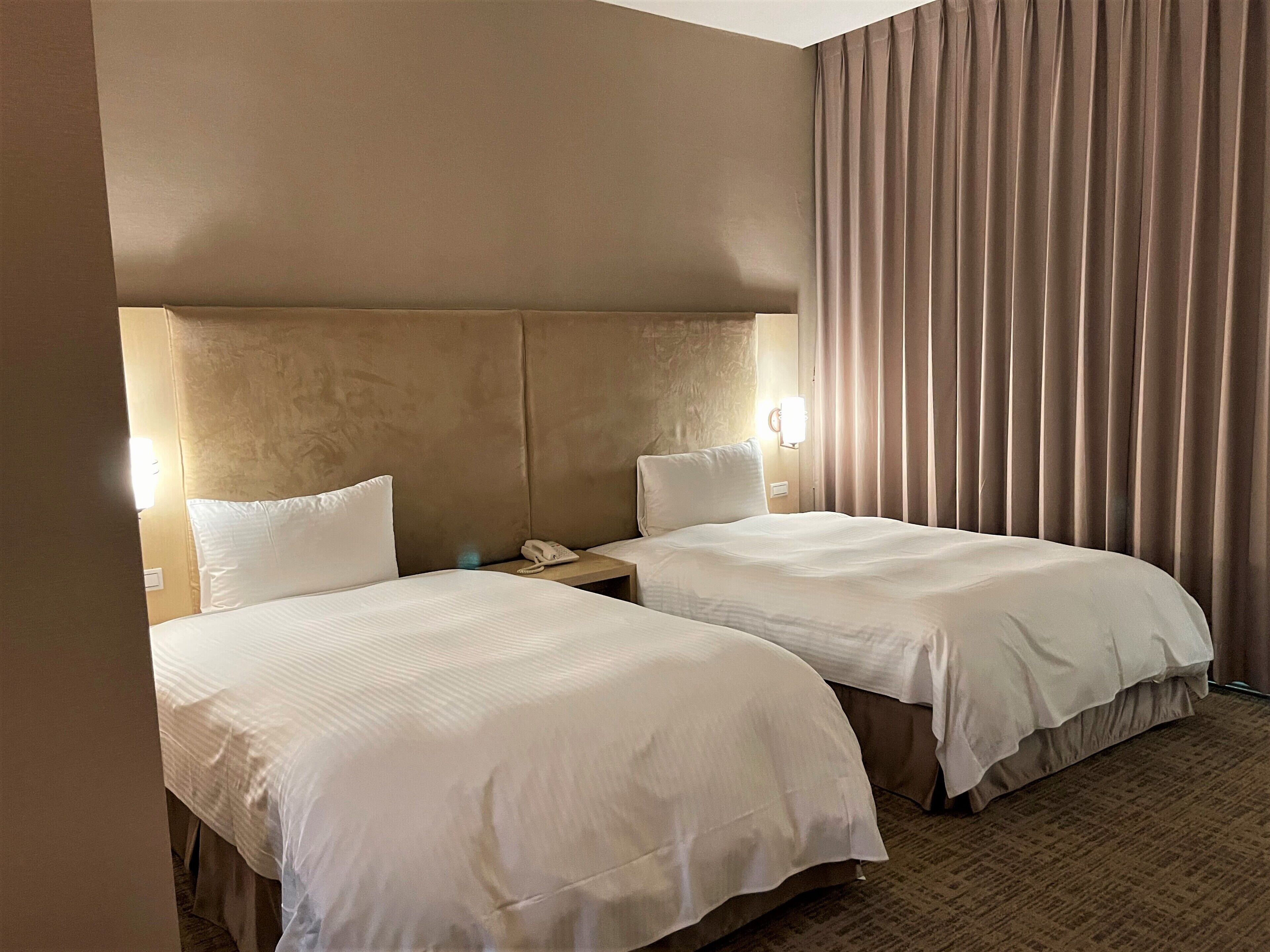 A hotel room with two beds, a headboard, a nightstand with a phone, and a window with curtains.