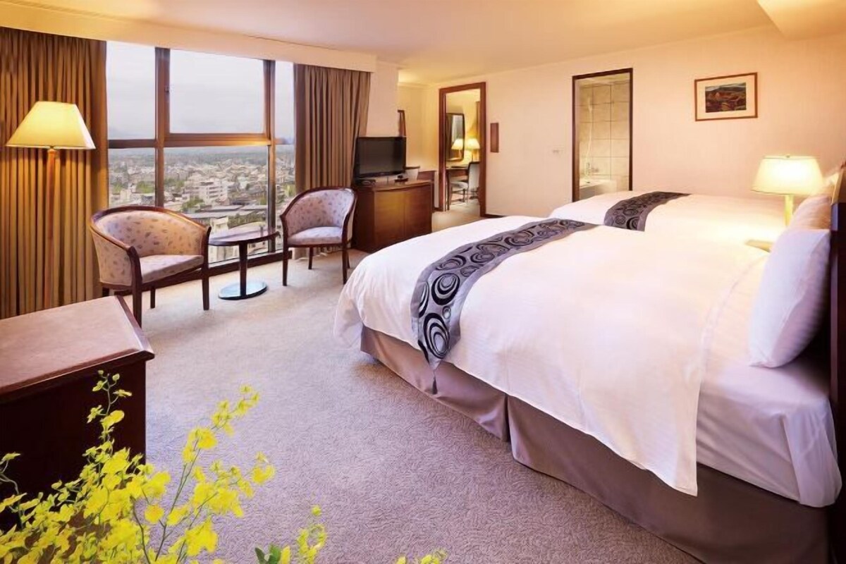 Habitación de hotel con una cama grande, un escritorio, una silla, un televisor y vistas de la ciudad a través de amplias ventanas.