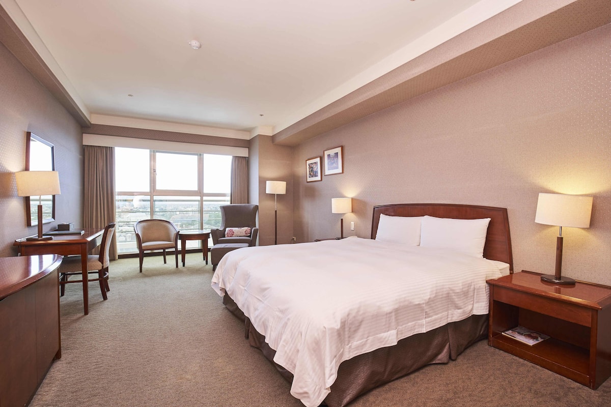A hotel room with a large bed, a desk, a chair, and a nightstand with a lamp.