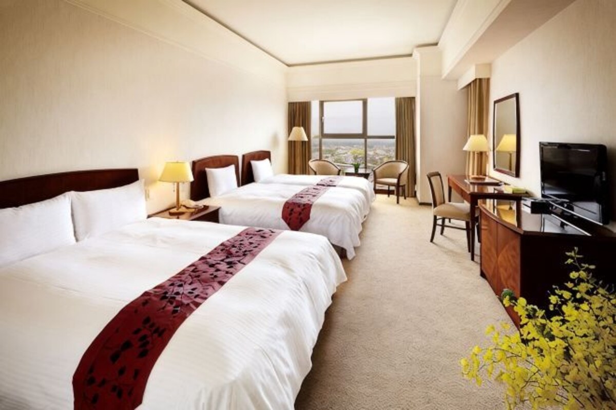 Habitación de hotel con dos camas, un escritorio, una silla, televisión y vistas a la ciudad.
