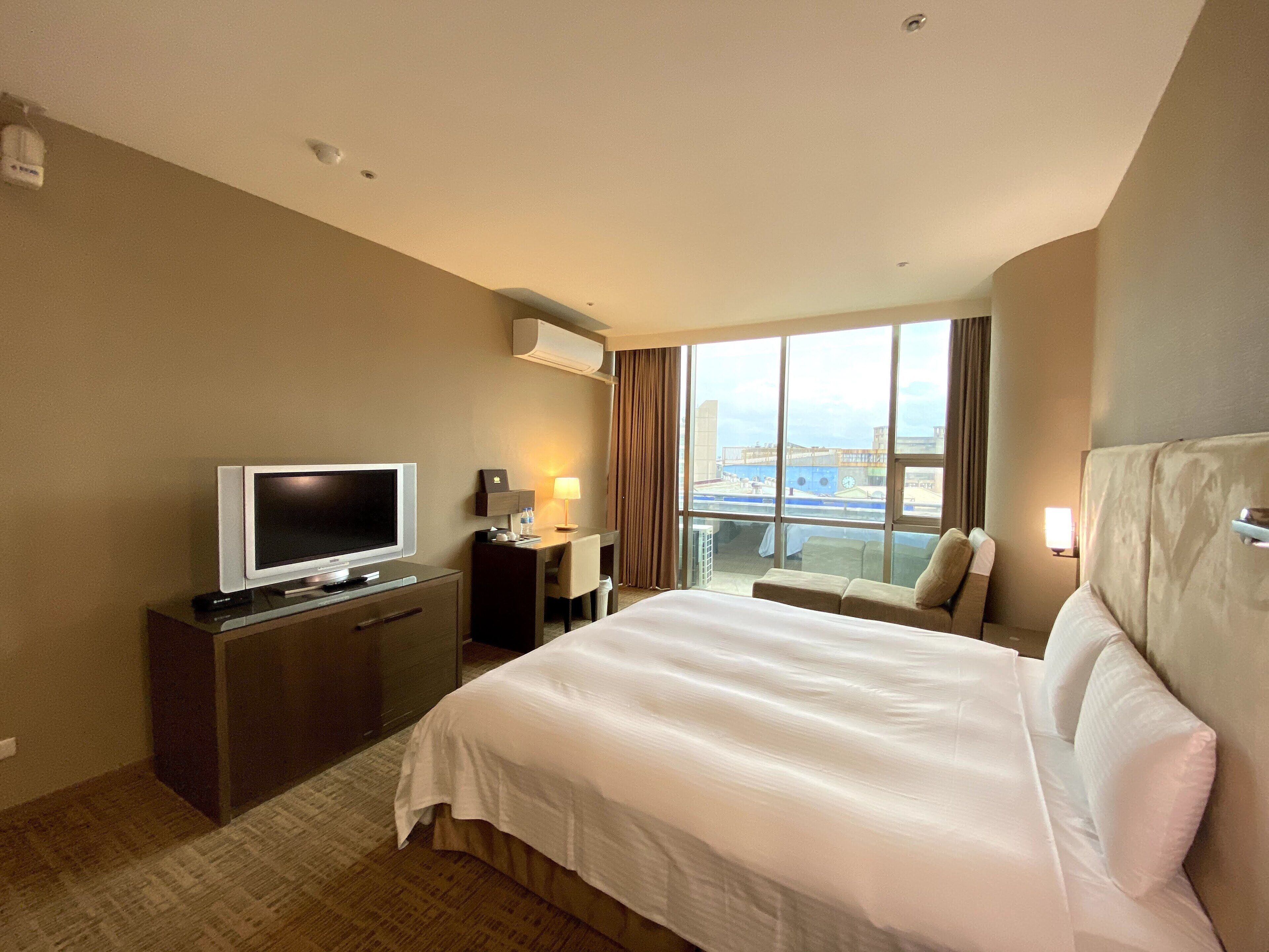 A hotel room with a bed, a desk with a TV, a window with a view, and a ceiling with recessed lighting.