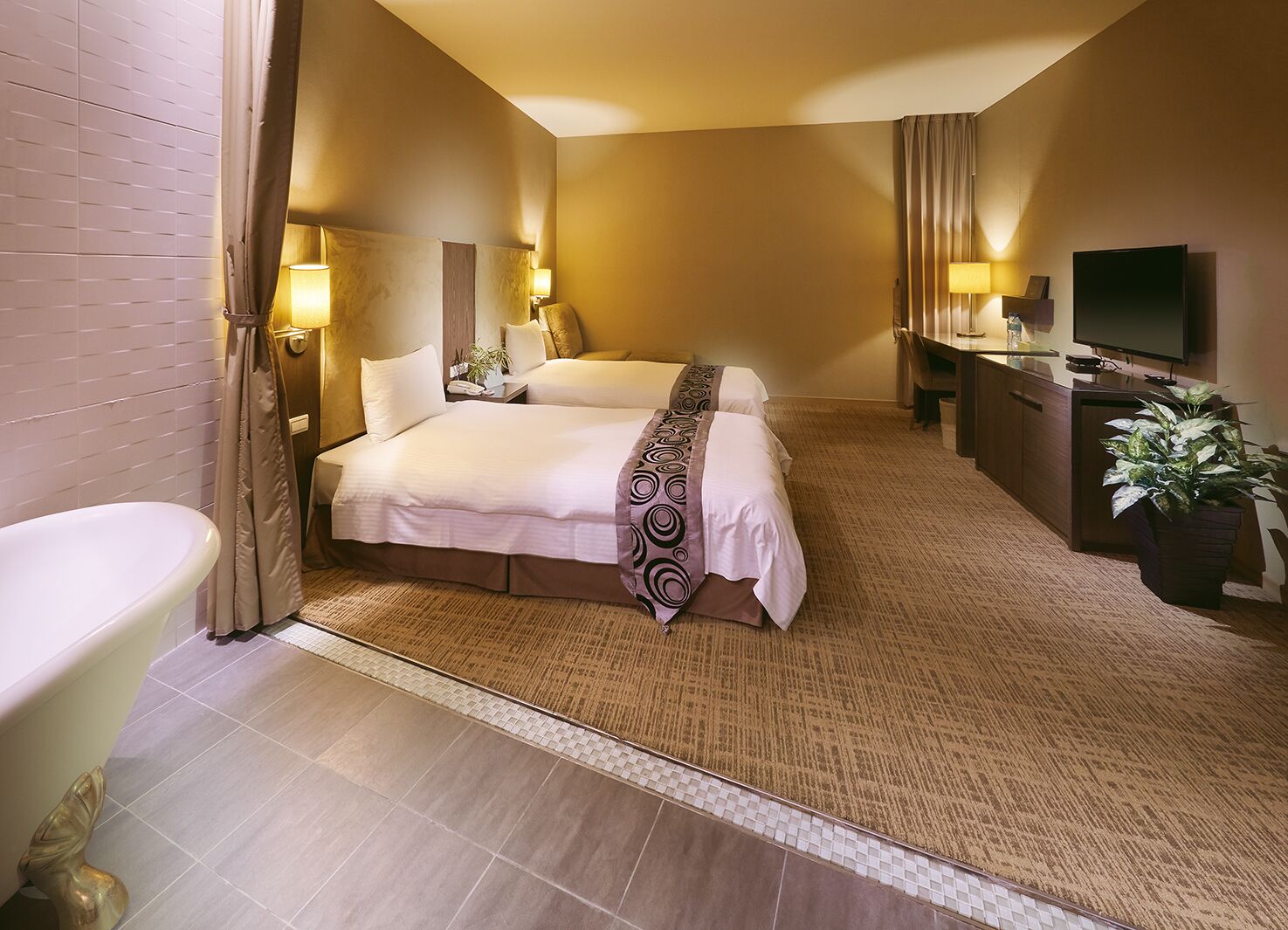 A hotel room with two beds, a bathroom, a desk with a TV, and a potted plant.
