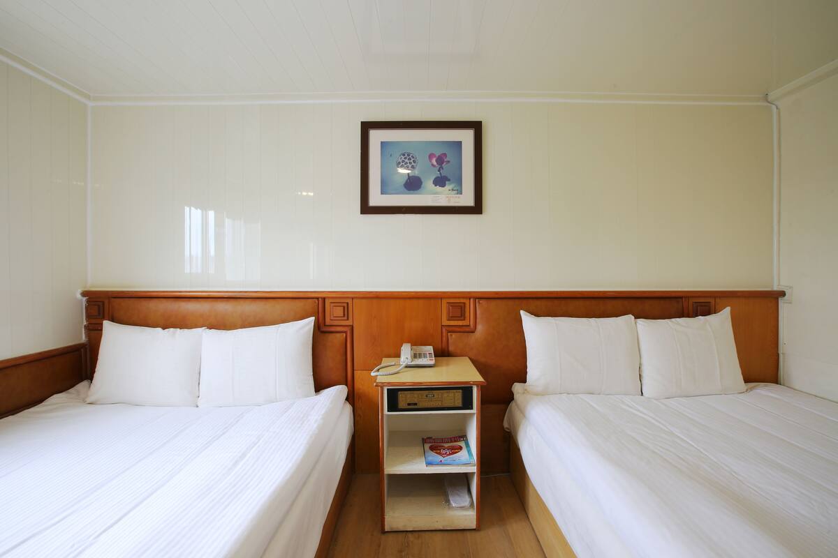 A hotel room with two beds, a wooden headboard, a bedside table with a phone and a book, and a framed picture on the wall.