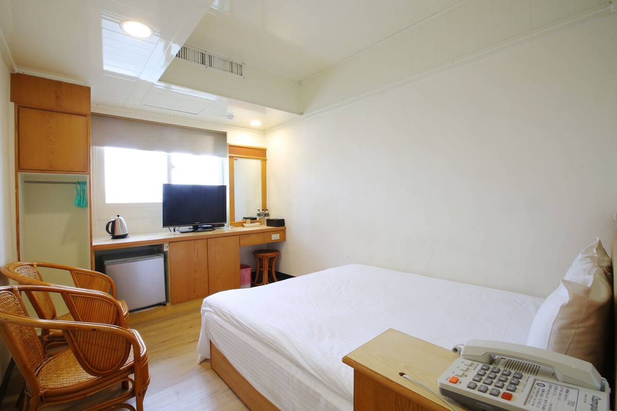 A hotel room with a bed, a desk with a television, a telephone, and wicker chairs.