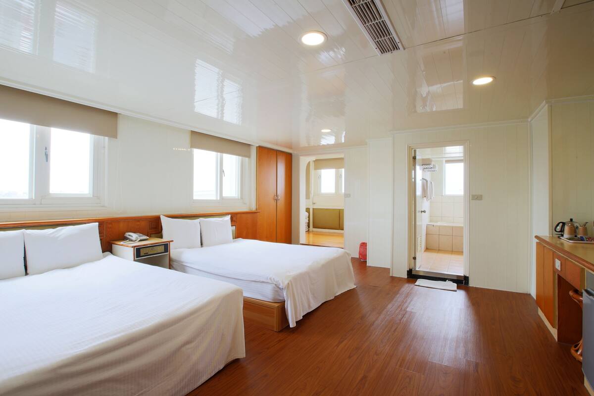 A hotel room with two beds, wooden flooring, a small desk with a telephone, and a view of the bathroom through an open door.