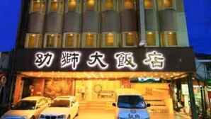Un hotel con fachada moderna, señalización iluminada y un estacionamiento con varios autos.