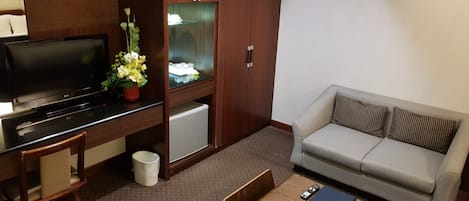 A hotel room with a TV, a small fridge, a sofa, a wooden coffee table, and a desk with a chair.