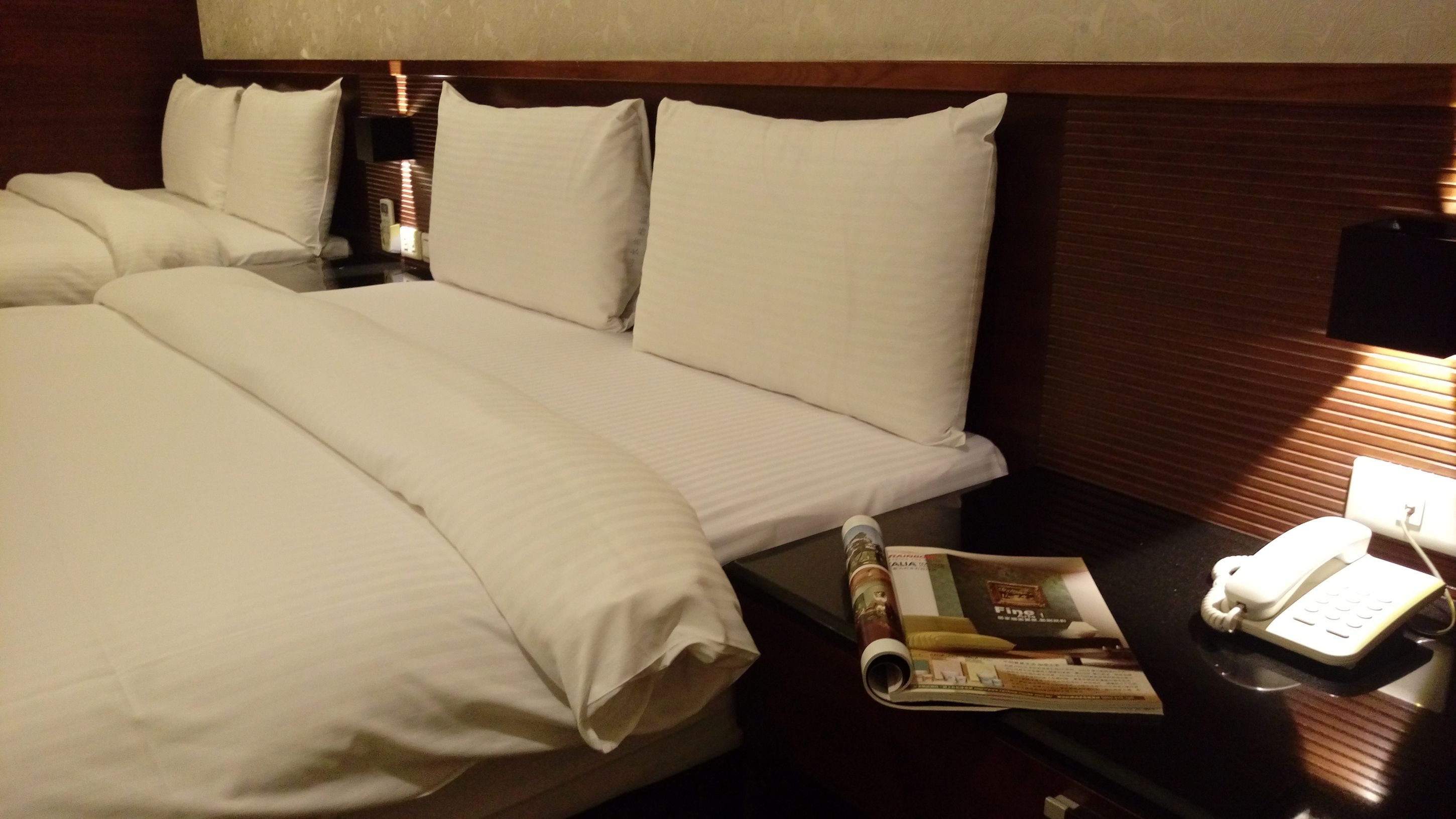 Una habitación de hotel con una cama grande, una mesita de noche con una revista, un teléfono y una lámpara.