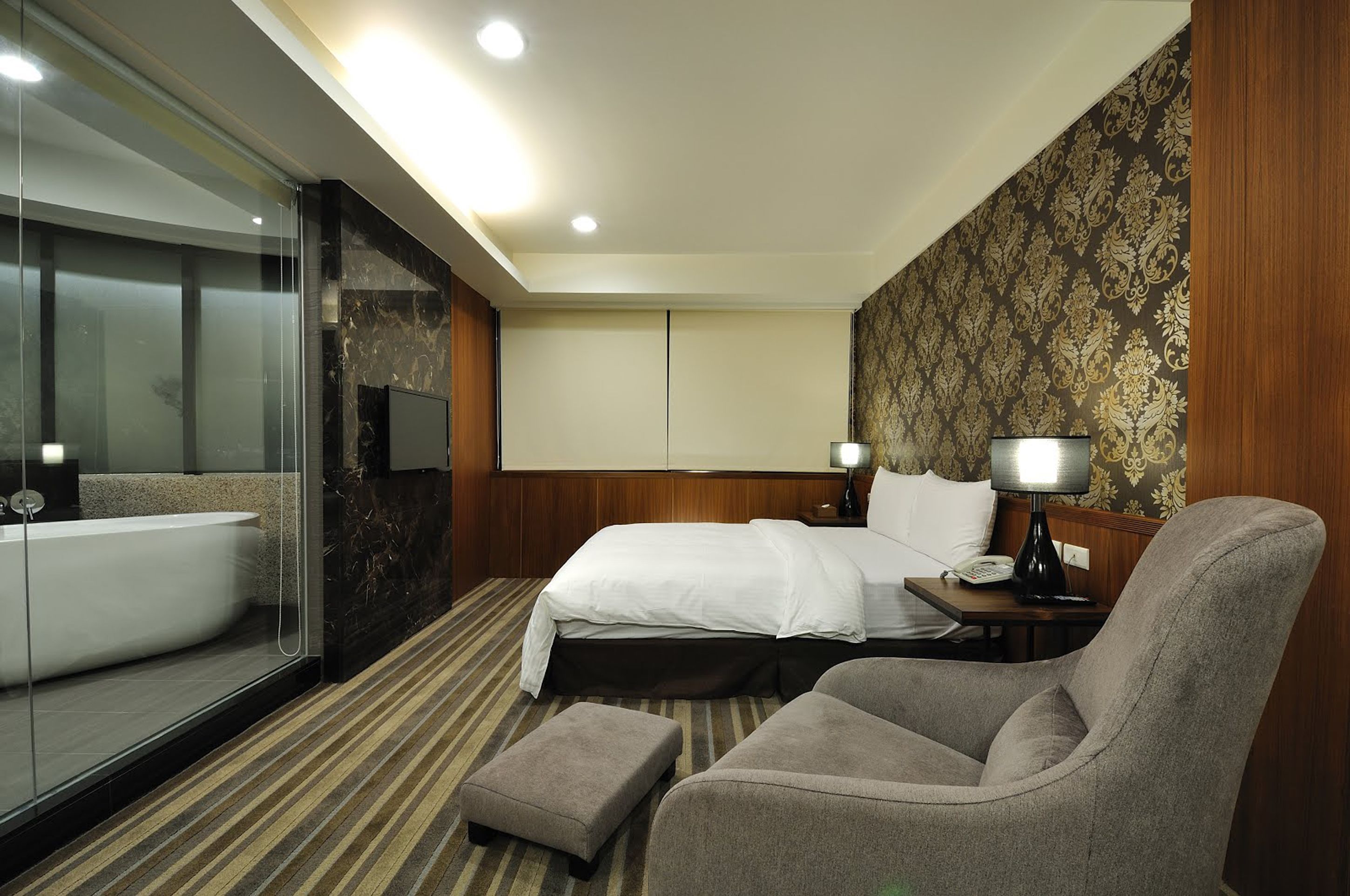 A hotel room with a large bed, a sitting area with a chair and ottoman, a glass-enclosed shower, and a wooden headboard.