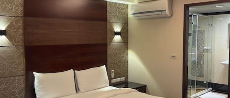 Habitación de hotel con una cama grande, cabecera de madera, aire acondicionado de pared y mampara de ducha de vidrio.