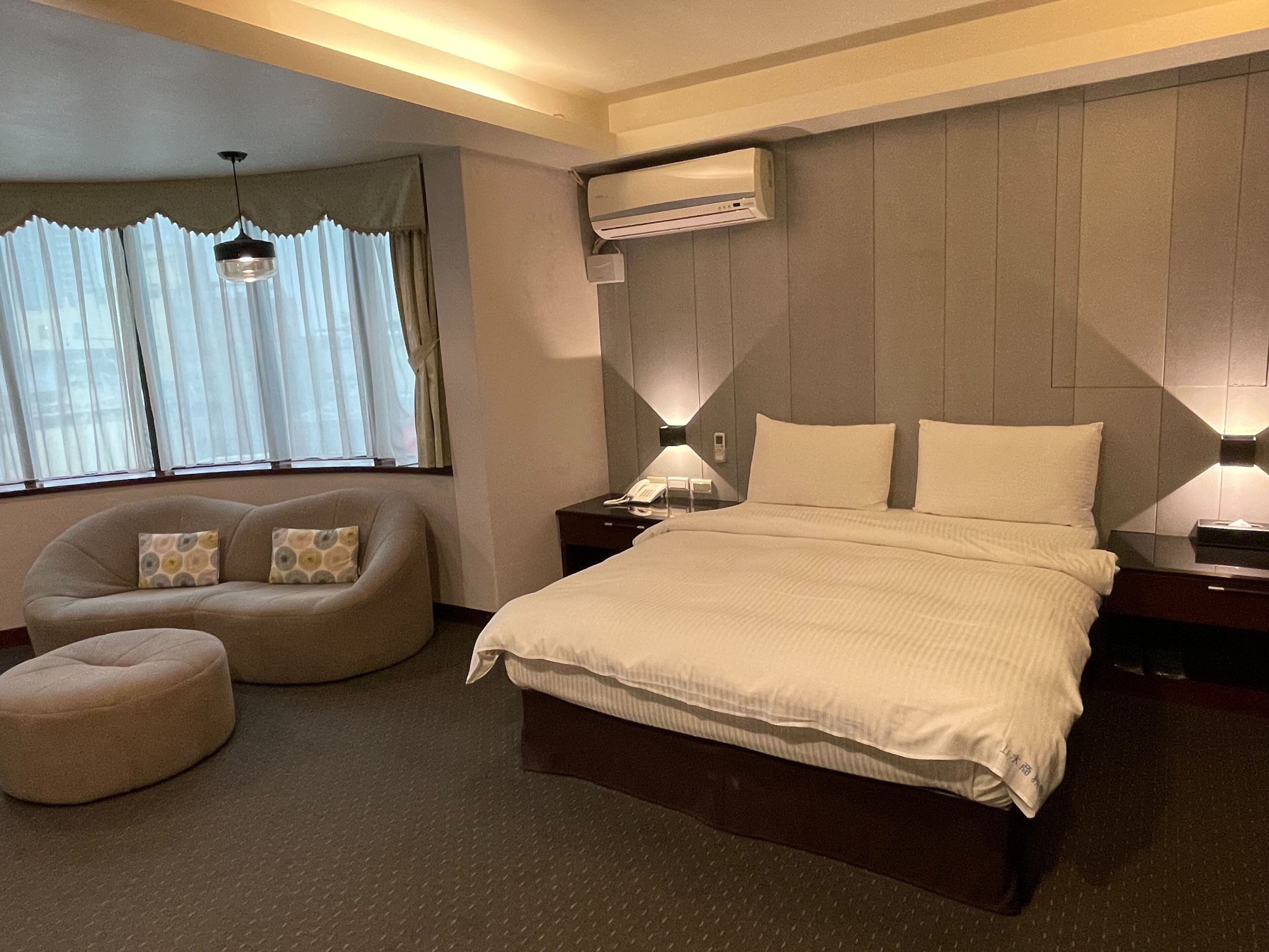 Una habitación de hotel con una cama grande, un sofá, un pequeño reposapiés, una mesita de noche, una lámpara y un equipo de aire acondicionado.