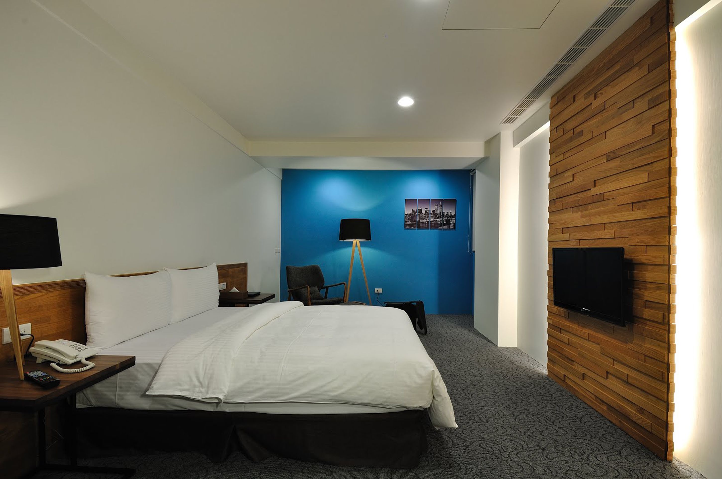 Habitación de hotel con una cama grande, un escritorio con teléfono, una silla, una lámpara de escritorio, un televisor de pantalla plana montado en una pared de madera y una pared azul decorada con una imagen de un paisaje urbano.