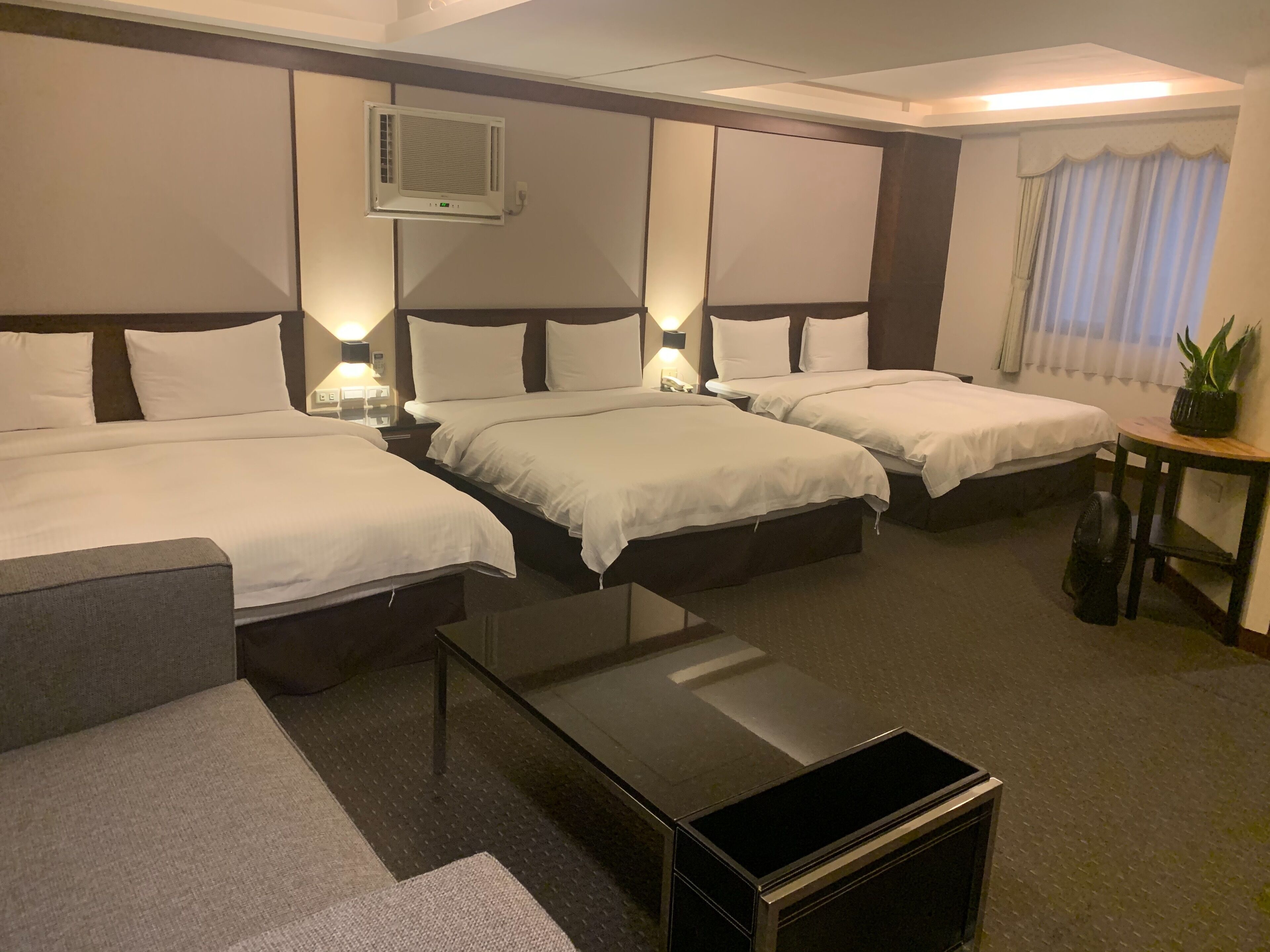 Hotelkamer met twee bedden, een bureau, een stoel, een klein tafeltje met een plant en een staande ventilator.