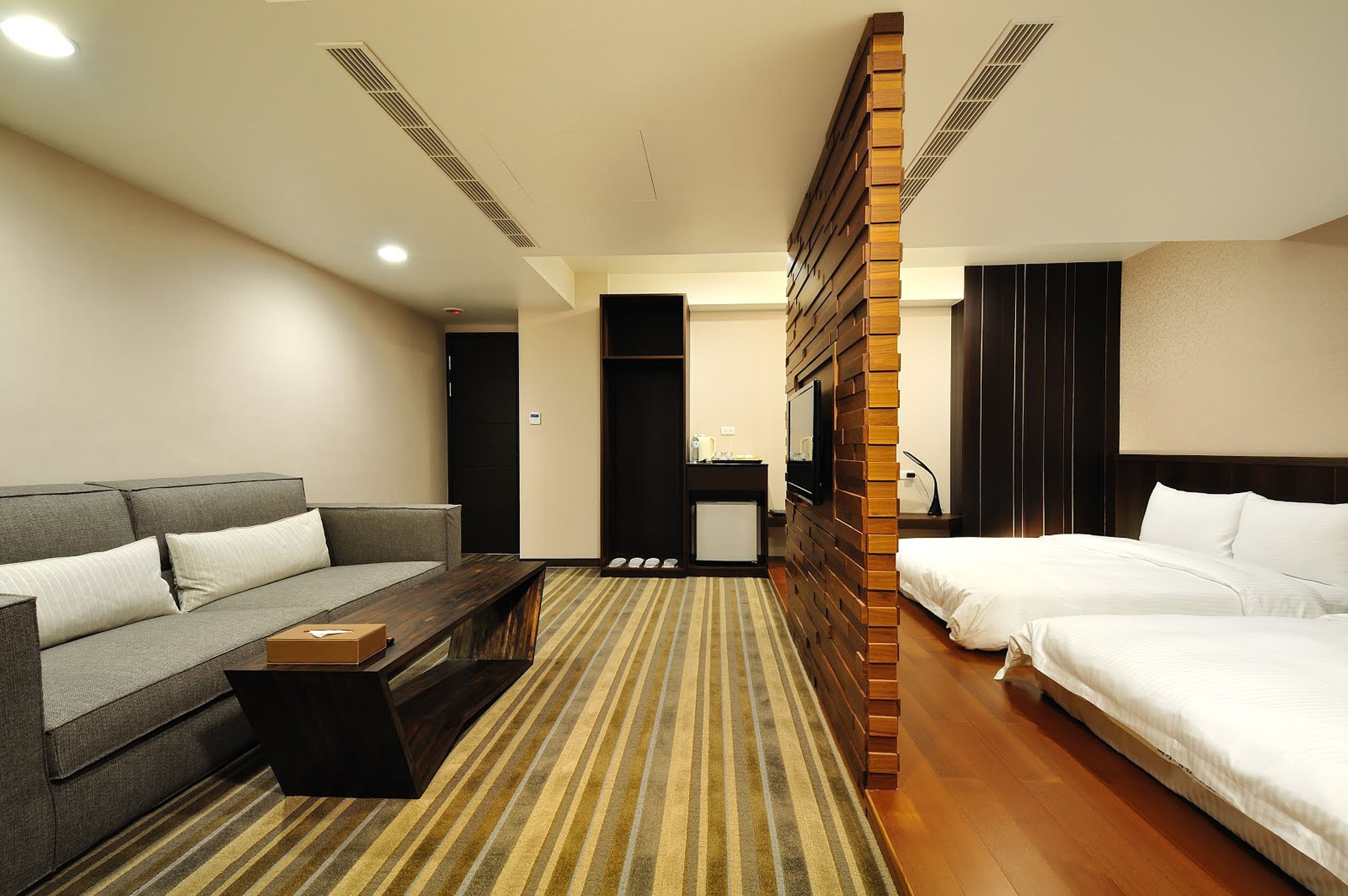 A hotel room with a sofa, a bed, a wooden headboard, a small table, and a striped carpet.