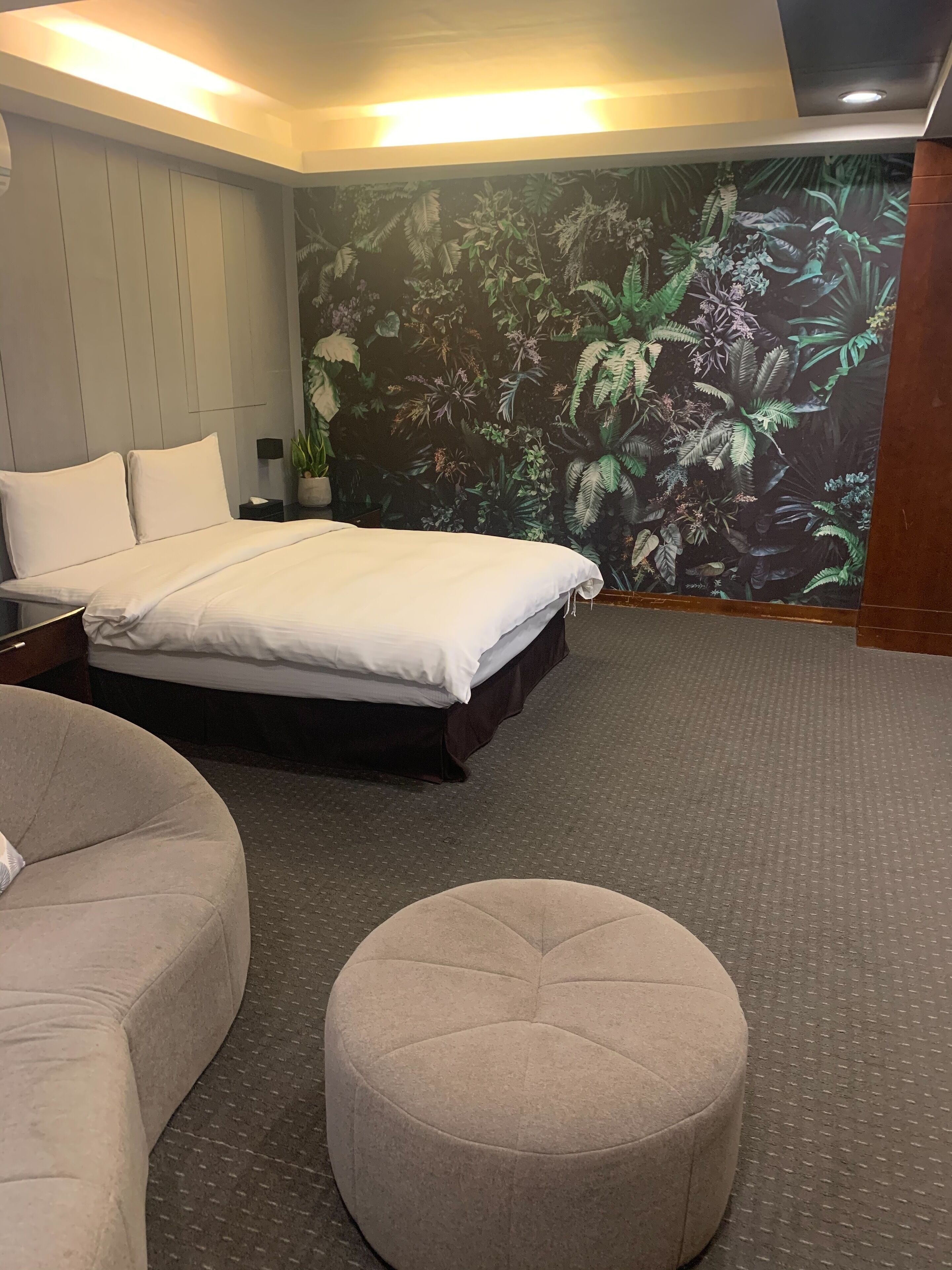 Ein Schlafzimmer mit einem Bett, einem runden Hocker, einem Nachttisch und einer Wand mit einem tropischen Wandbild.