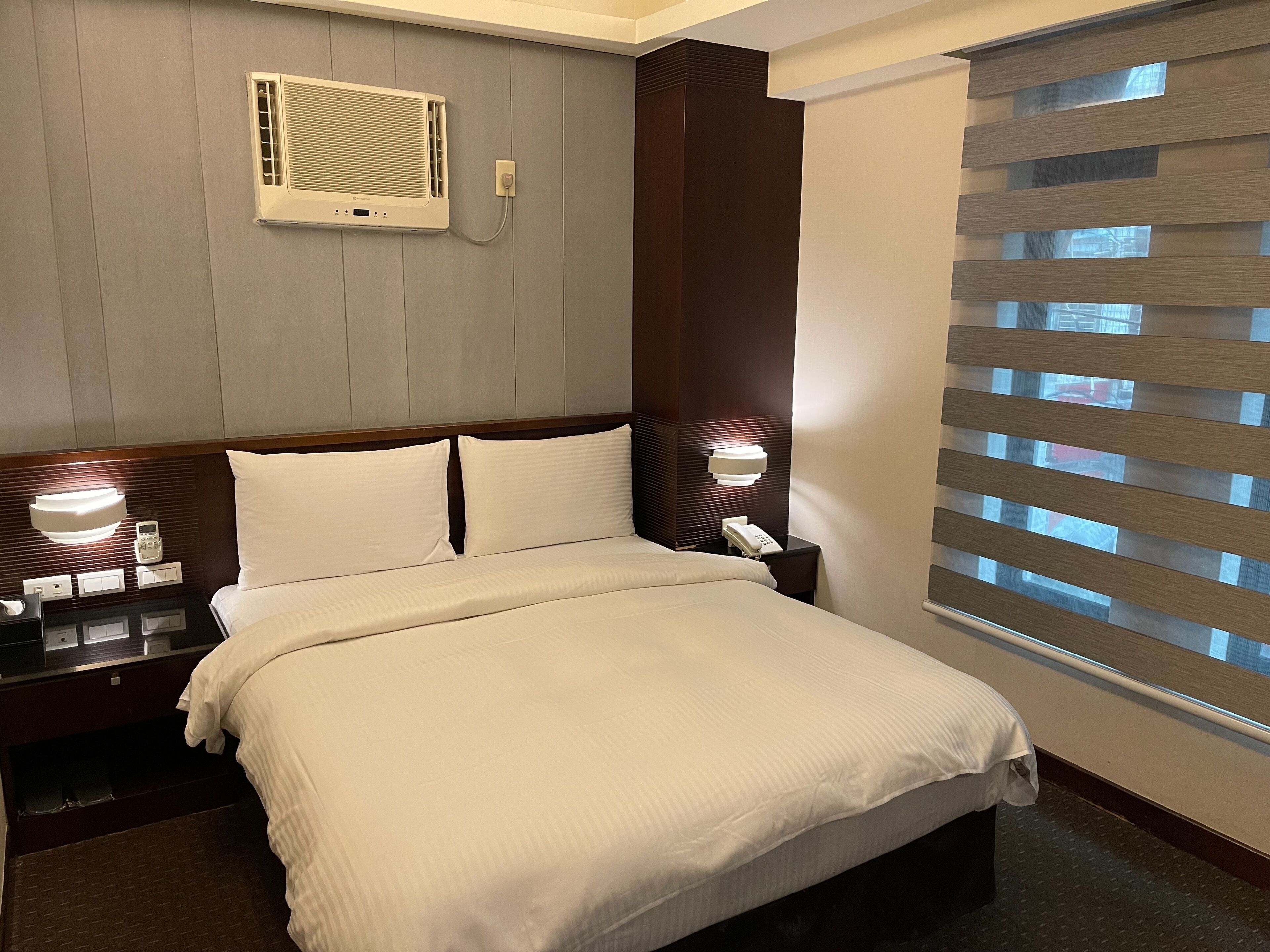 Habitación de hotel con cama, mesitas de noche, teléfono, lámpara y ventana con persianas.