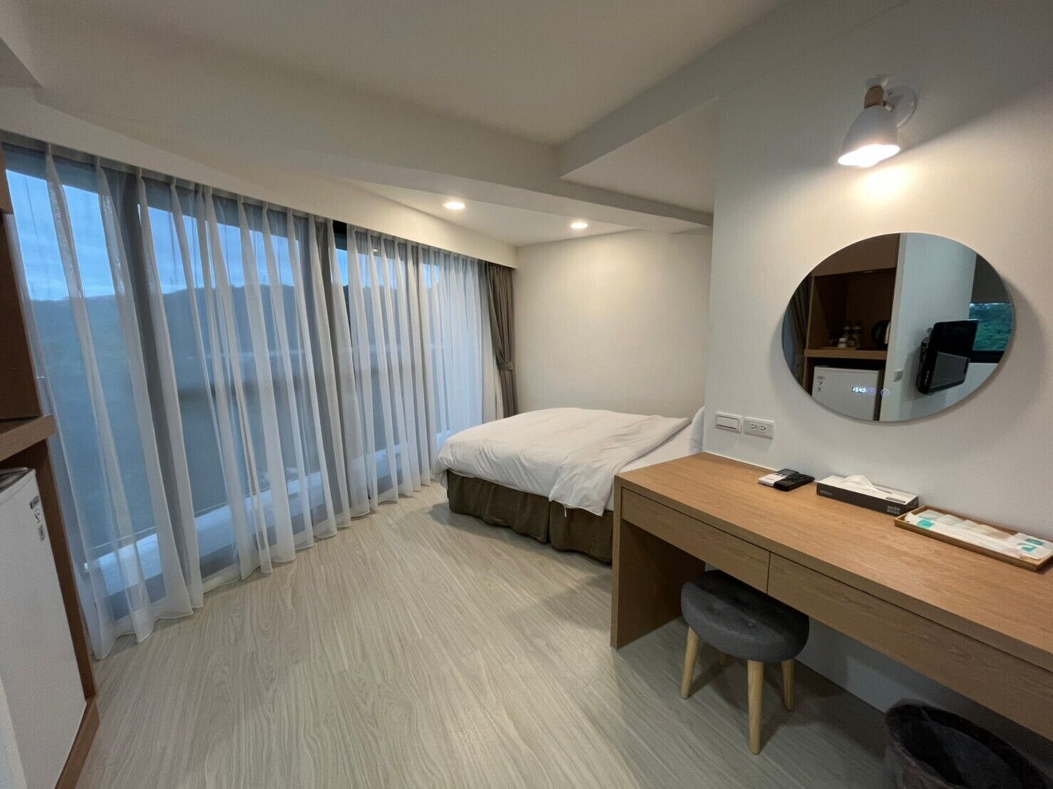 Habitación de hotel con cama, escritorio, silla, espejo redondo y ventana con cortinas traslúcidas.