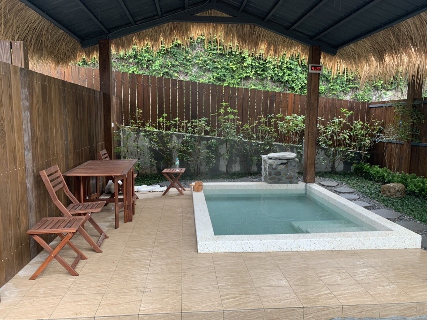 A covered patio with a pool, wooden furniture, and a bamboo fence.
