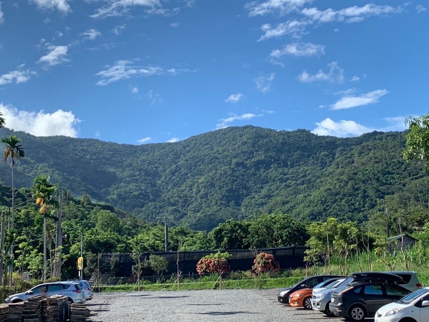 Zona de estacionamiento con coches, superficie de grava y una montaña verde exuberante al fondo bajo un cielo despejado.