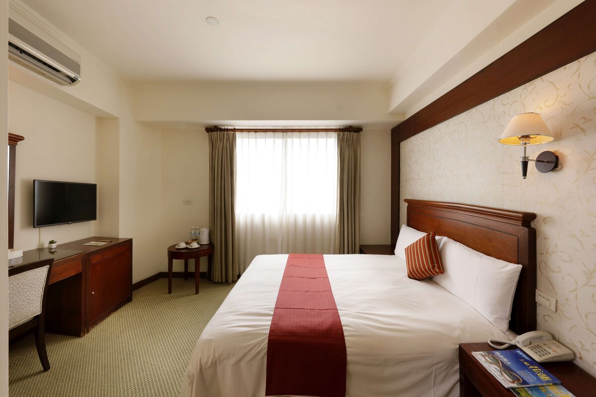 Habitación de hotel con cama, escritorio, televisión de pantalla plana, teléfono y ventana con cortinas.