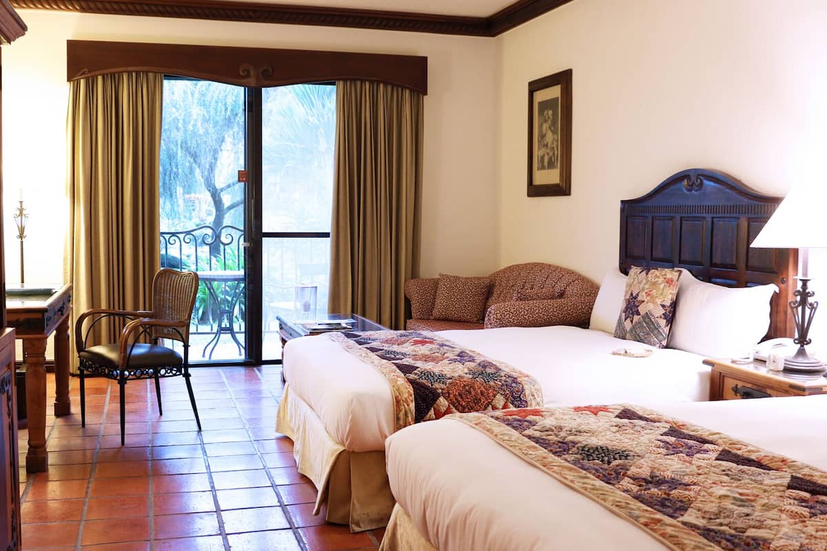 A hotel room with two beds, a television, a desk with a chair, and a balcony with curtains.