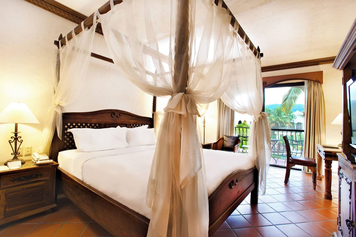 A four-poster bed with a canopy, a bedside table with a lamp, and a view of the outdoors through a window.