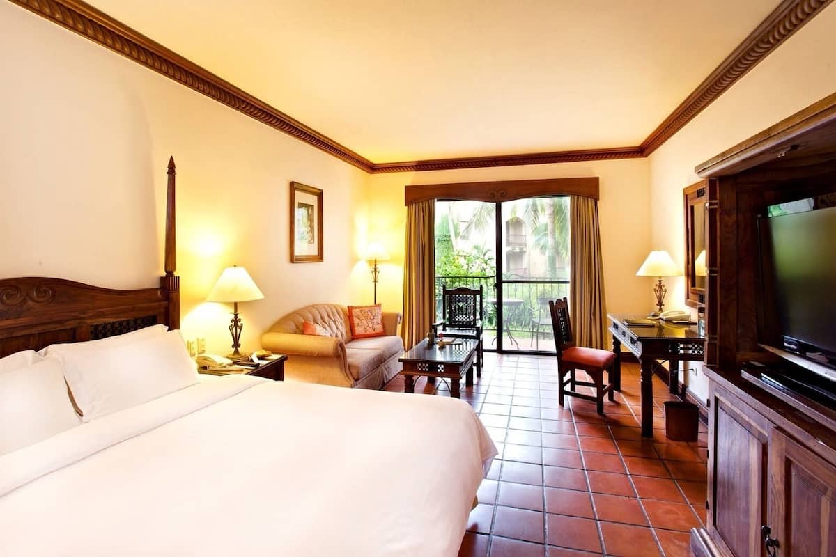 A hotel room with a large bed, a desk, a chair, a television, and a balcony with a view of greenery.