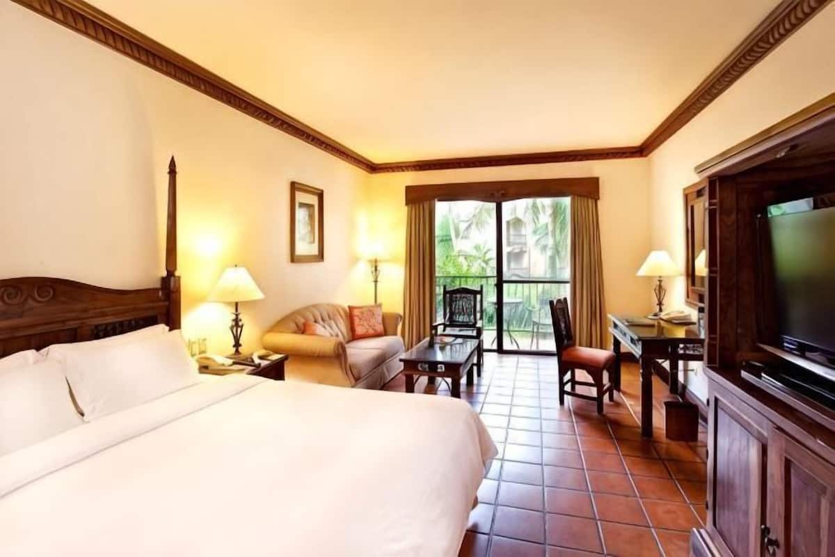 A hotel room with a large bed, a desk with a chair, a television, and a balcony with a view.