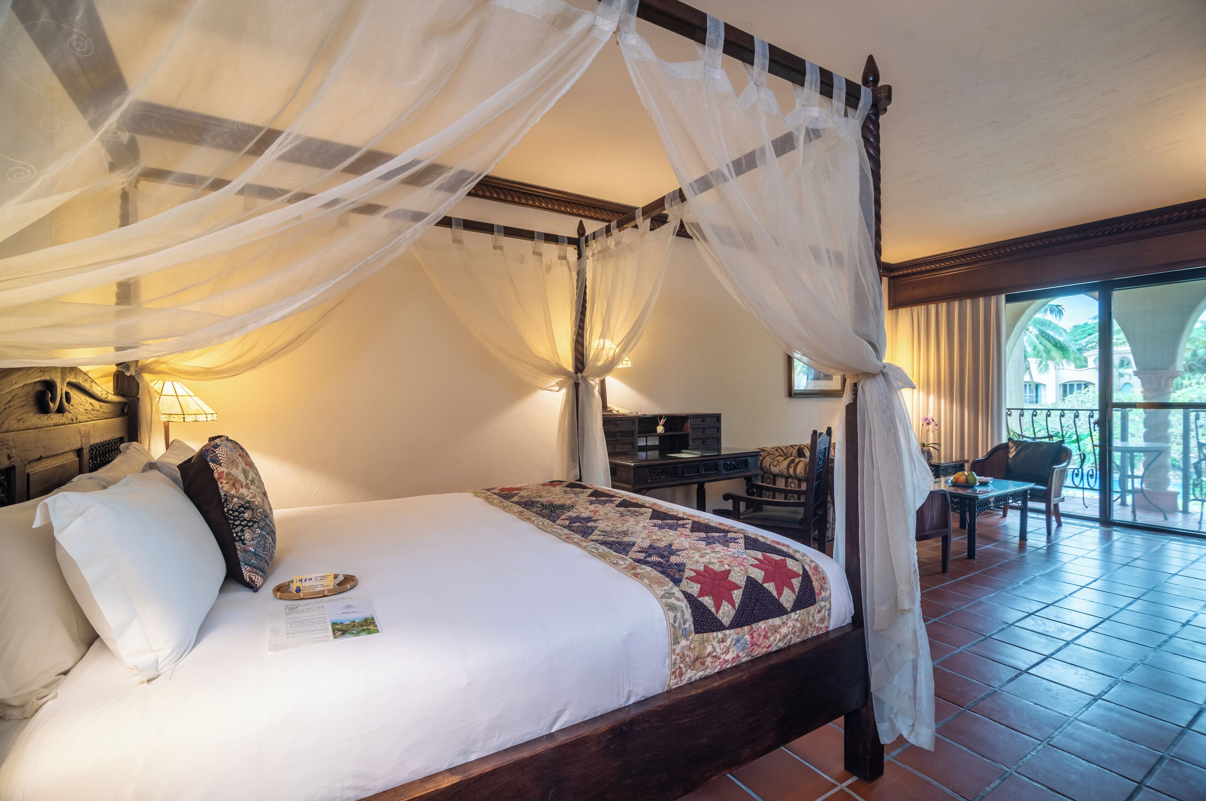 A four-poster bed with a canopy, a desk, and chairs in a room with a view of palm trees.