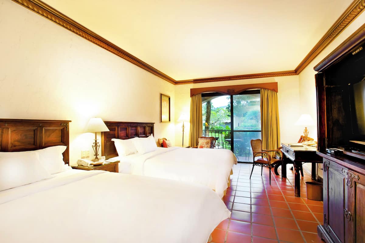 A hotel room with two beds, a desk, a chair, a television, and a balcony with a view of greenery.