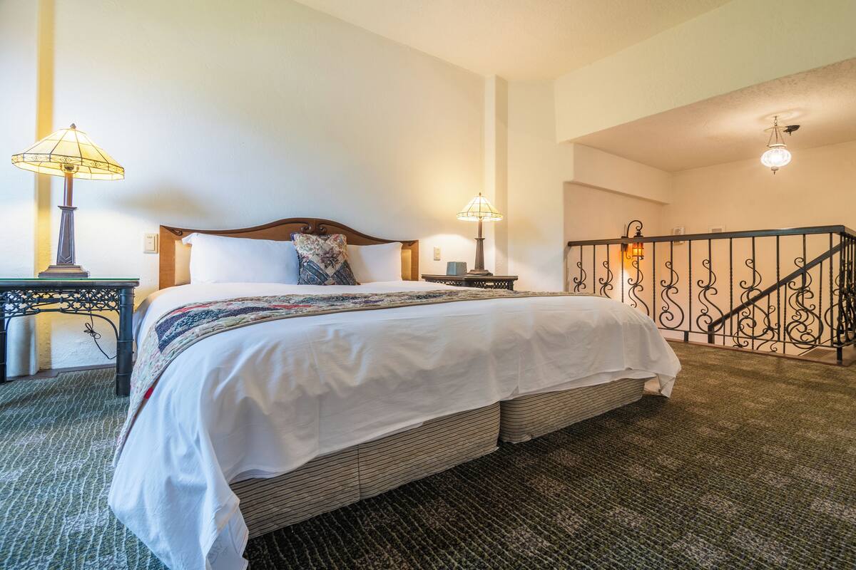A neatly made bed with a white comforter and pillows, a bedside table with a lamp, and a staircase with a wrought iron railing.