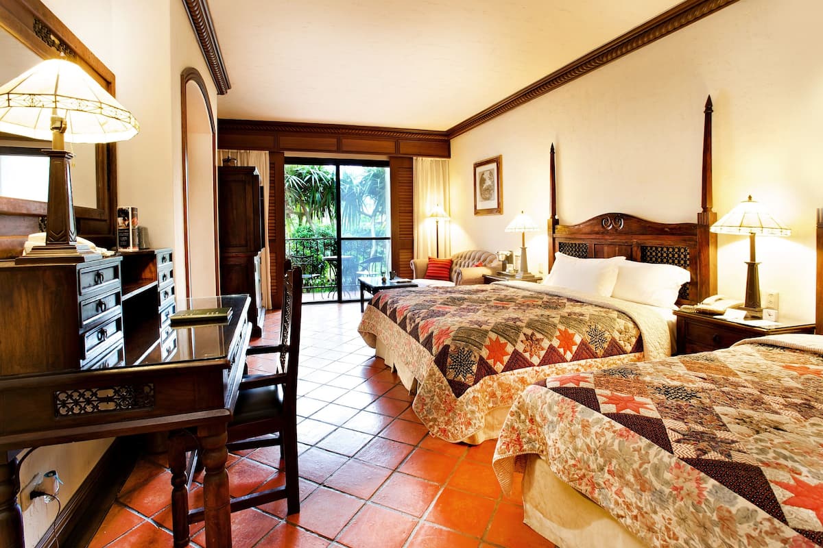 A hotel room with two beds, a desk with a lamp, a chair, and a view of the outdoors.