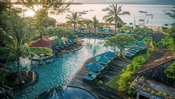 Zwembad van een resort met ligbedden en parasols, omgeven door palmbomen en met uitzicht op een kalme zee met boten.