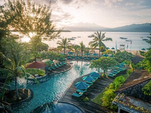 A resort pool with lounge chairs and umbrellas, surrounded by palm trees and overlooking a calm sea with boats.