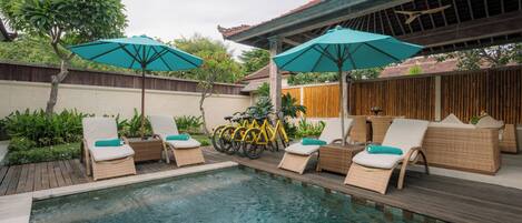 A pool area with lounge chairs and umbrellas, surrounded by wooden fences and greenery.