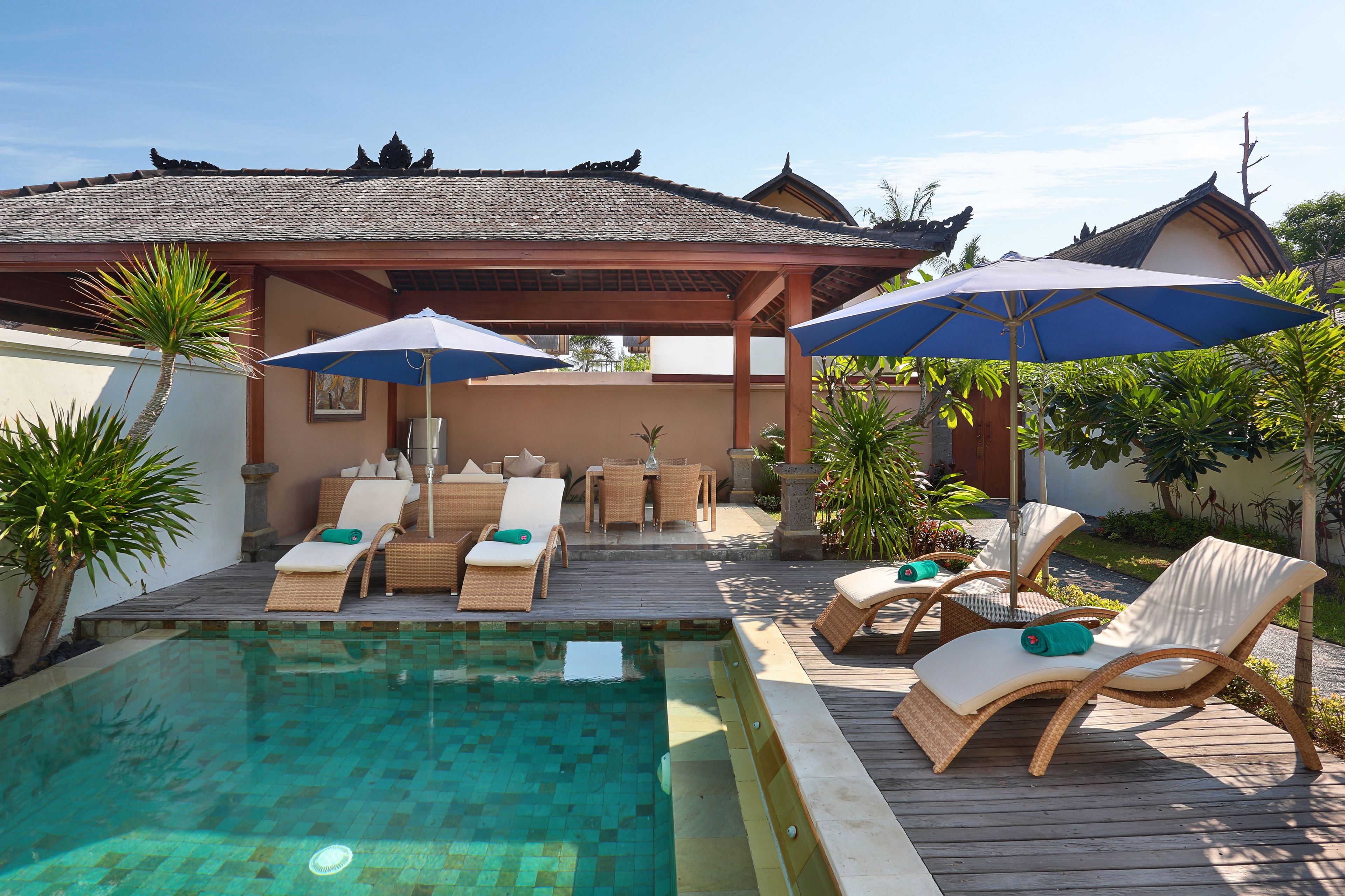 Uma área de piscina com espreguiçadeiras e guarda-sóis, rodeada por um deck de madeira e plantas tropicais.