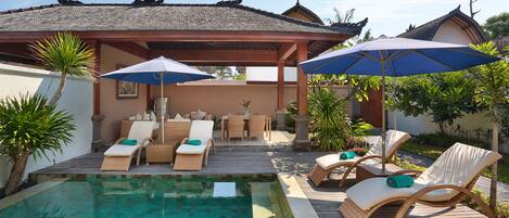 Uma área de piscina com espreguiçadeiras e guarda-sóis, rodeada por um deck de madeira e plantas tropicais.