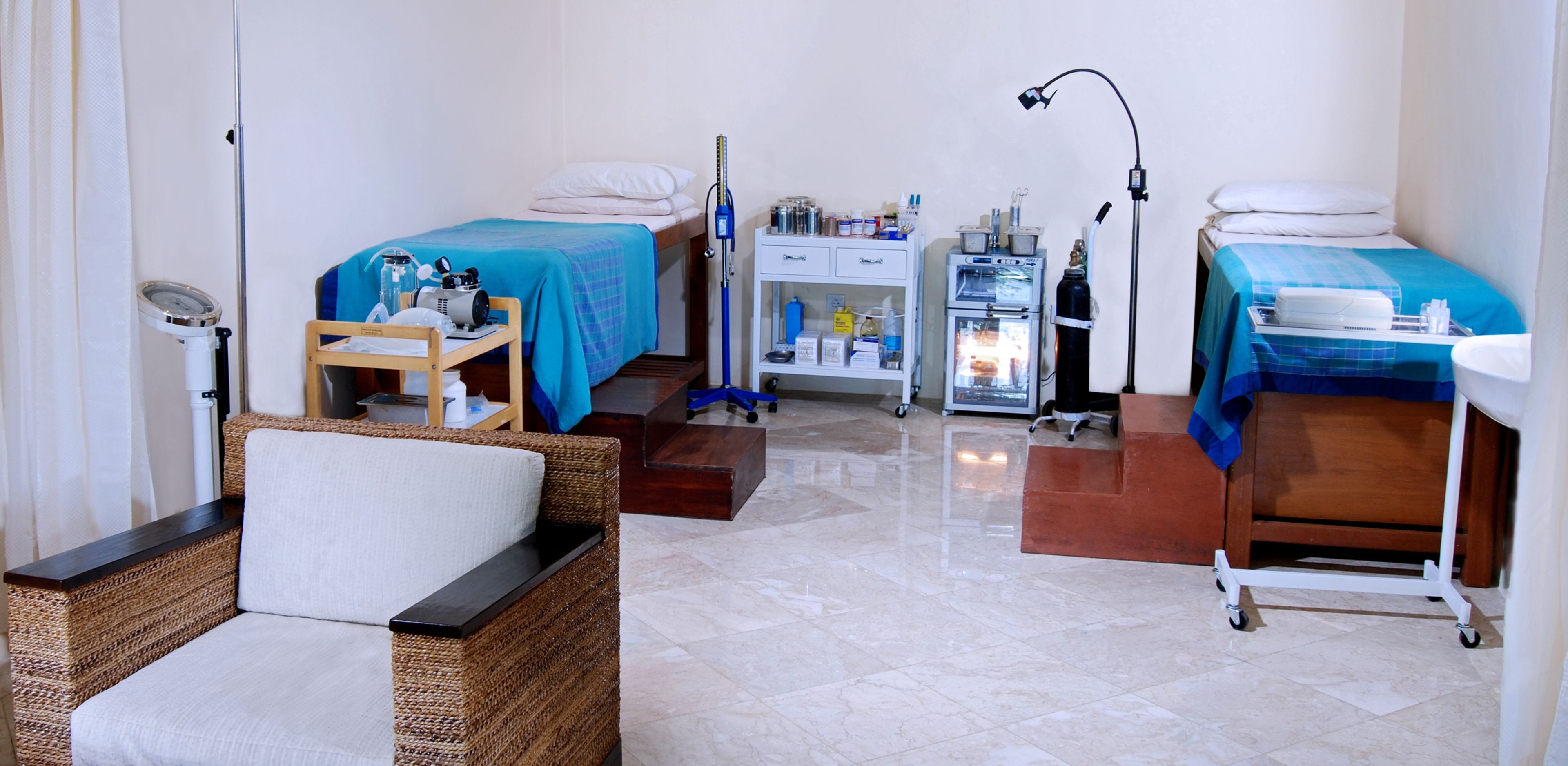 Una habitación con dos camas, una mesita pequeña con una silla, un carrito con suministros médicos y un refrigerador.