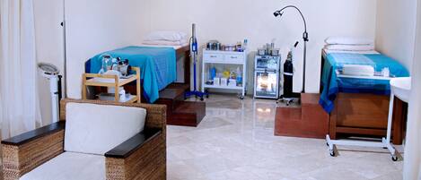 Una habitación con dos camas, una mesita pequeña con una silla, un carrito con suministros médicos y un refrigerador.
