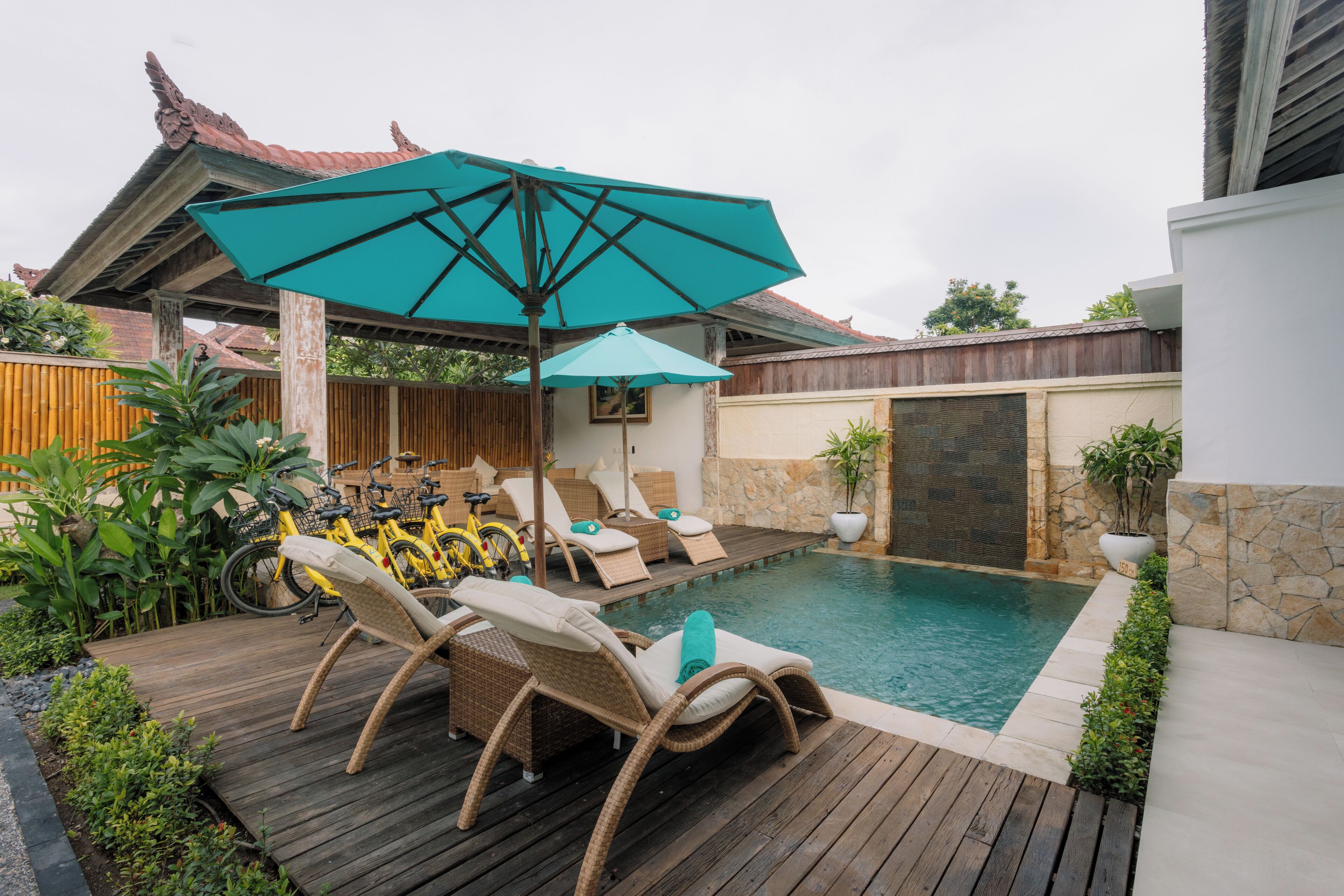 Zona de piscina con sillones de descanso y una mesa, rodeada de plantas y una terraza de madera.