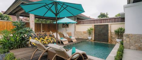 Zona de piscina con sillones de descanso y una mesa, rodeada de plantas y una terraza de madera.