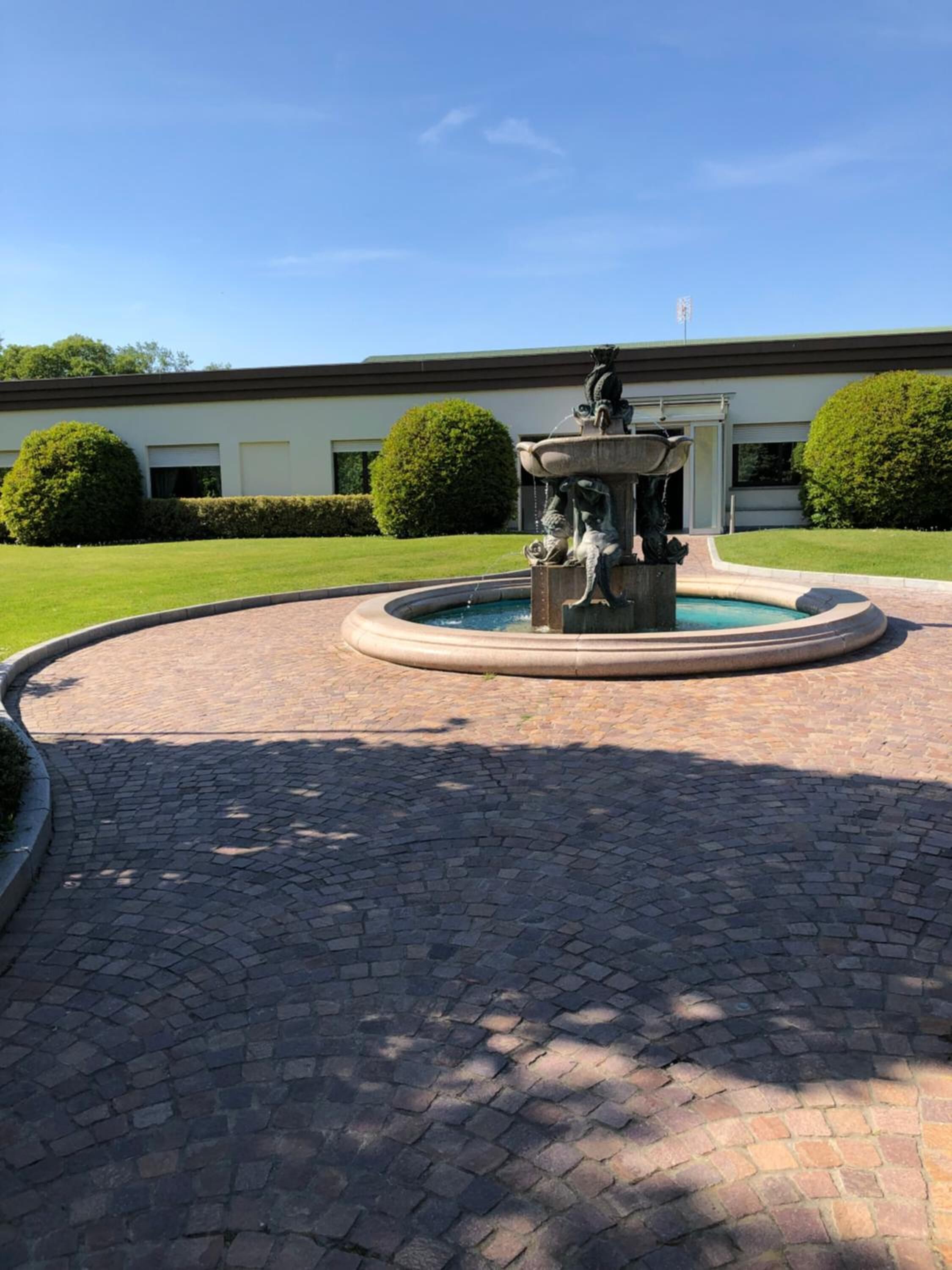 Un chemin circulaire pavé de briques mène à une fontaine ornée d’une statue au centre, le tout étant entouré de haies bien entretenues et d’un bâtiment visible au loin.