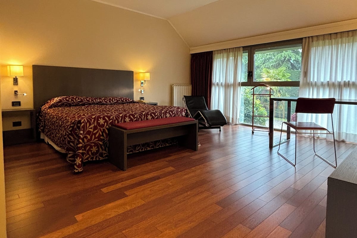 Amplia habitación con una cama grande, un escritorio y una silla, con vista a la vegetación por la ventana.